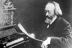 Photo en noir et blanc du compositeur Max Bruch en train de lire une partition devant un piano - Agrandir l'image